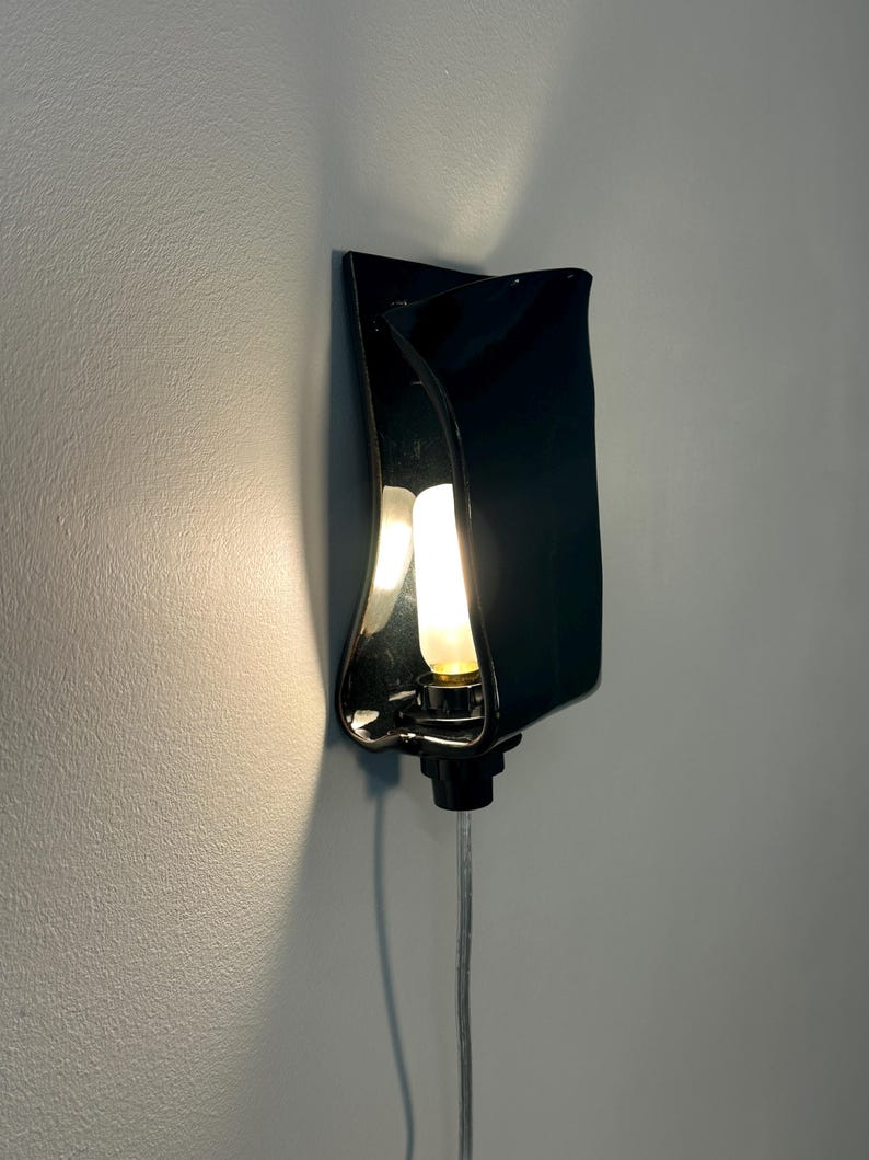 May include: A wall-mounted lamp featuring a glossy black ceramic shade with a curved, folded design. The fixture holds a single glowing light bulb in the center, casting a warm light against the wall, and includes a clear power cord extending downward.