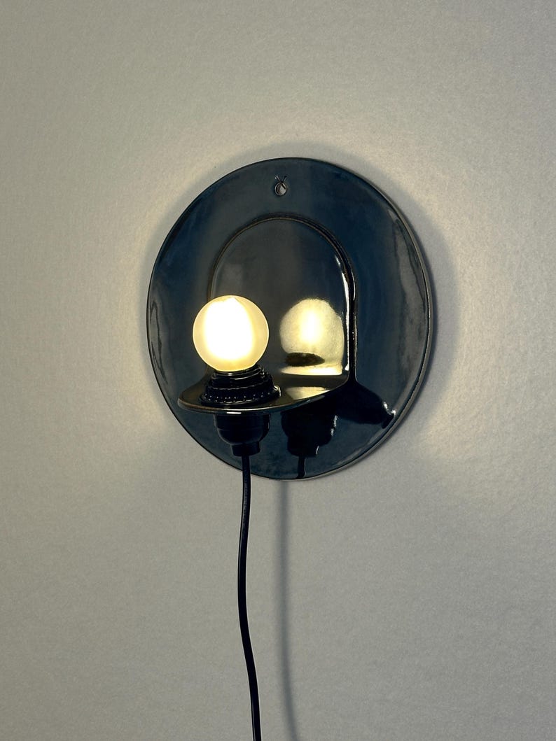 May include: A circular, glossy black ceramic wall sconce featuring a small, protruding shelf that holds a single glowing, spherical light bulb. A thin black power cord extends downward from the base of the light fixture, which is mounted flush.