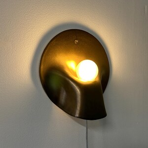 May include: A wall-mounted sconce featuring a dark bronze-toned, circular metal backplate with a fluid, organic, scooped-out shape. A single, glowing, spherical white light bulb is positioned within the recessed curve, emitting a warm light.