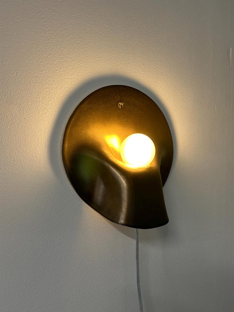 May include: A wall-mounted sconce featuring a dark bronze-toned, circular metal backplate with a fluid, organic, scooped-out shape. A single, glowing, spherical white light bulb is positioned within the recessed curve, emitting a warm light.