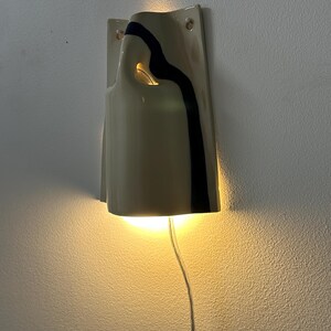 May include: A wall-mounted ceramic sconce with a glossy cream finish and a single vertical black stripe. The fixture features an open top and a cutout side, emitting a warm, downward-facing light. A clear power cord extends from the bottom of the unit.