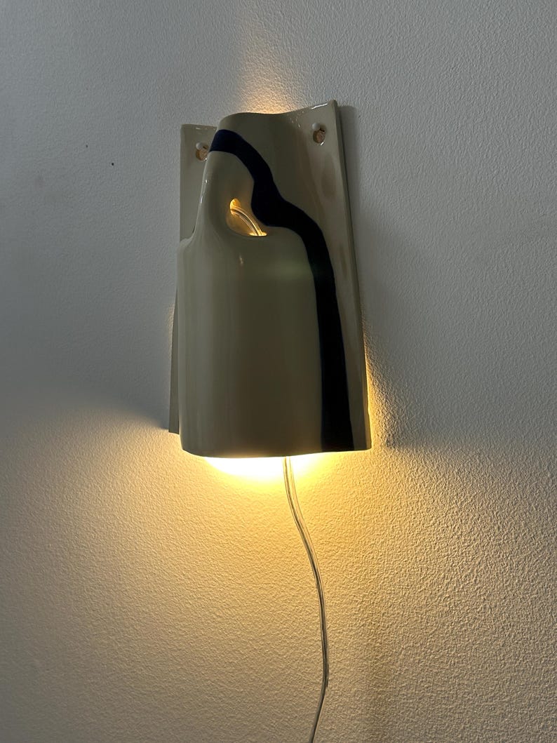 May include: A wall-mounted ceramic sconce with a glossy cream finish and a single vertical black stripe. The fixture features an open top and a cutout side, emitting a warm, downward-facing light. A clear power cord extends from the bottom of the unit.