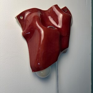 Puede incluir: Un aplique de pared de cer&aacute;mica con un acabado rojo intenso brillante y una superficie ondulada y plegada. La pieza cuenta con dos peque&ntilde;os orificios circulares para el montaje y una bombilla blanca que sobresale por la parte inferior con un cable.