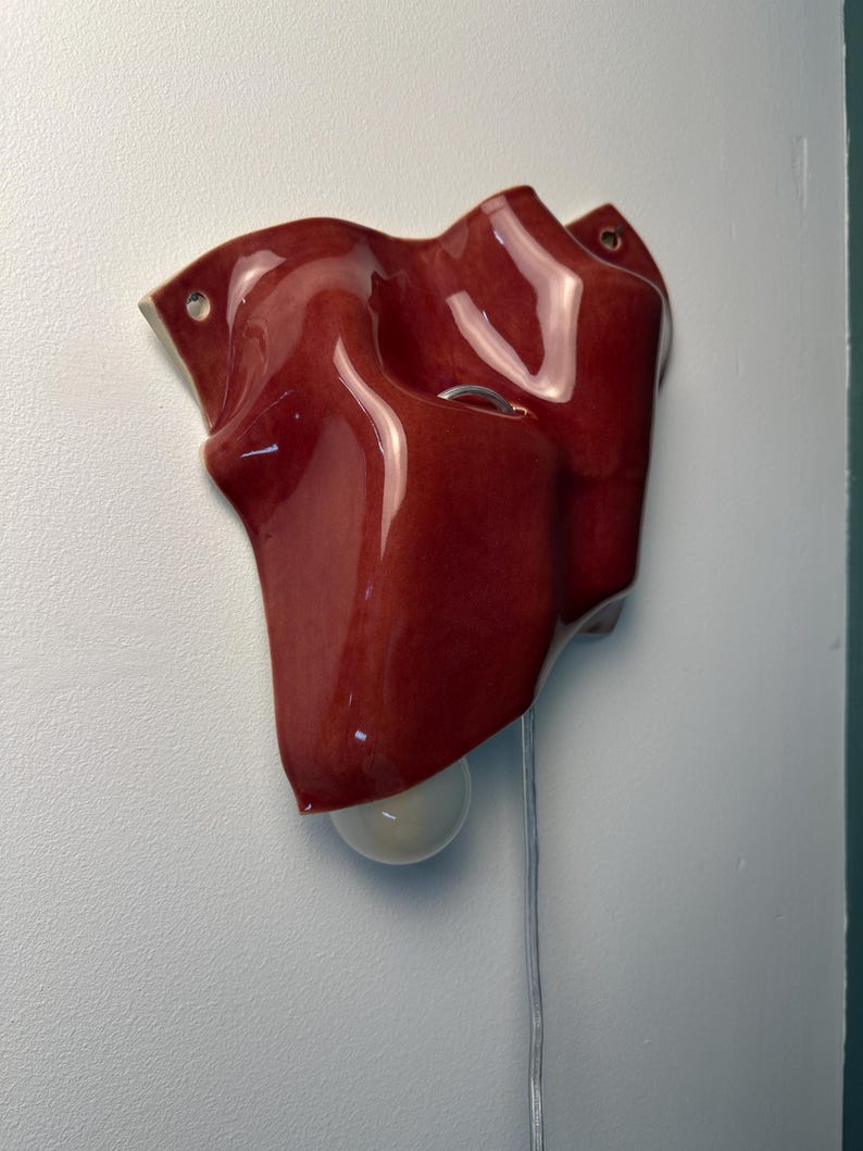 Puede incluir: Un aplique de pared de cer&aacute;mica con un acabado rojo intenso brillante y una superficie ondulada y plegada. La pieza cuenta con dos peque&ntilde;os orificios circulares para el montaje y una bombilla blanca que sobresale por la parte inferior con un cable.