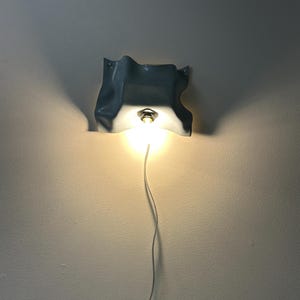 May include: A wall-mounted light fixture featuring a light blue, wavy, fabric-like ceramic shade with a central metallic socket holding a glowing light bulb. A thin white power cord extends downward from the base of the illuminated light fixture.