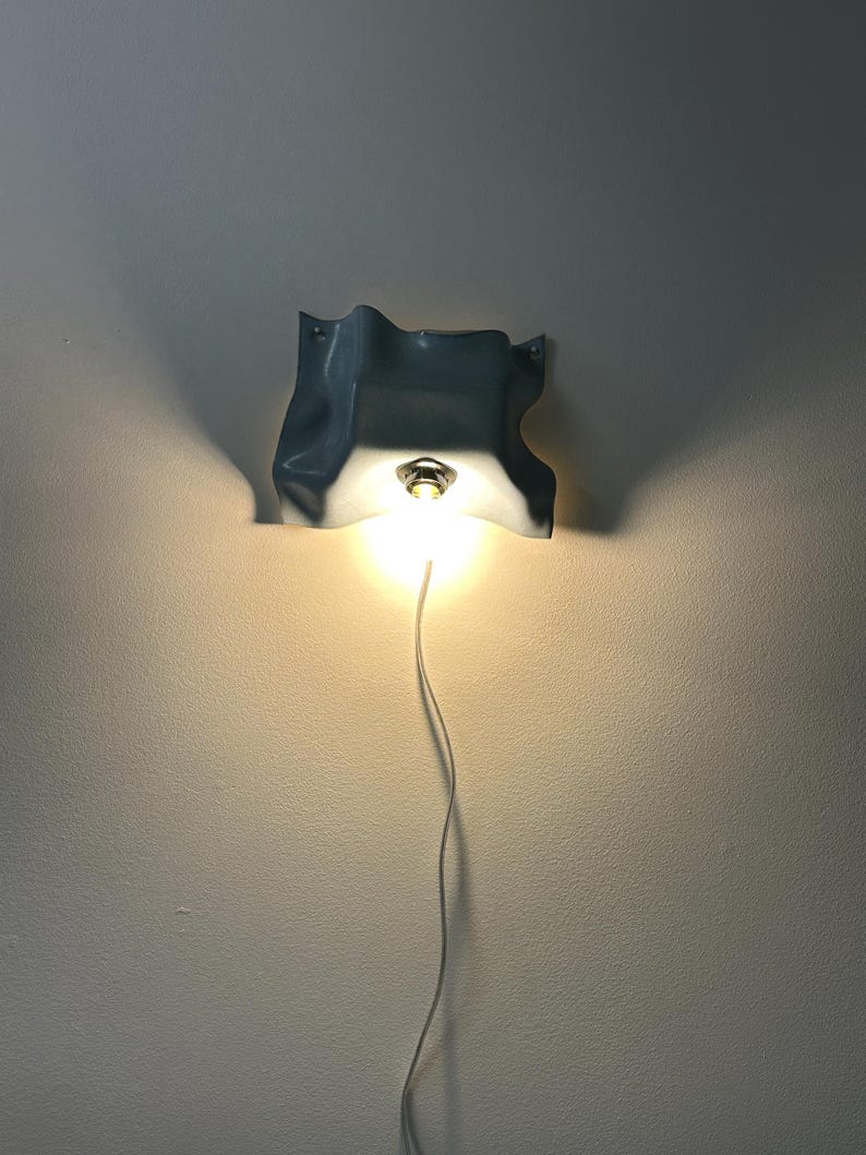 May include: A wall-mounted light fixture featuring a light blue, wavy, fabric-like ceramic shade with a central metallic socket holding a glowing light bulb. A thin white power cord extends downward from the base of the illuminated light fixture.
