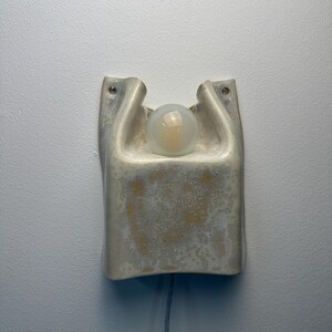 May include: A wall-mounted ceramic sconce featuring a textured, off-white glaze with subtle beige mottling. The fixture has a curved, organic shape with two upper mounting holes and a central circular opening that holds a single frosted glass bulb.