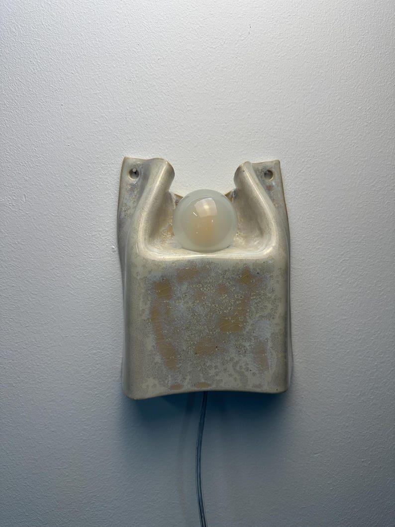May include: A wall-mounted ceramic sconce featuring a textured, off-white glaze with subtle beige mottling. The fixture has a curved, organic shape with two upper mounting holes and a central circular opening that holds a single frosted glass bulb.