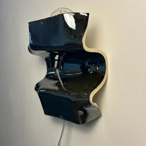 May include: A wall-mounted lamp featuring a glossy black ceramic body with a wavy, sculptural silhouette. The fixture includes a central black socket holding a clear light bulb and a transparent power cord extending from the bottom of the piece.