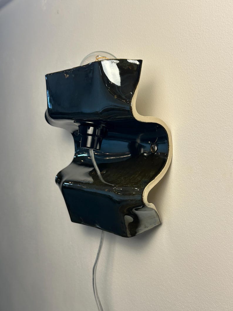 May include: A wall-mounted lamp featuring a glossy black ceramic body with a wavy, sculptural silhouette. The fixture includes a central black socket holding a clear light bulb and a transparent power cord extending from the bottom of the piece.