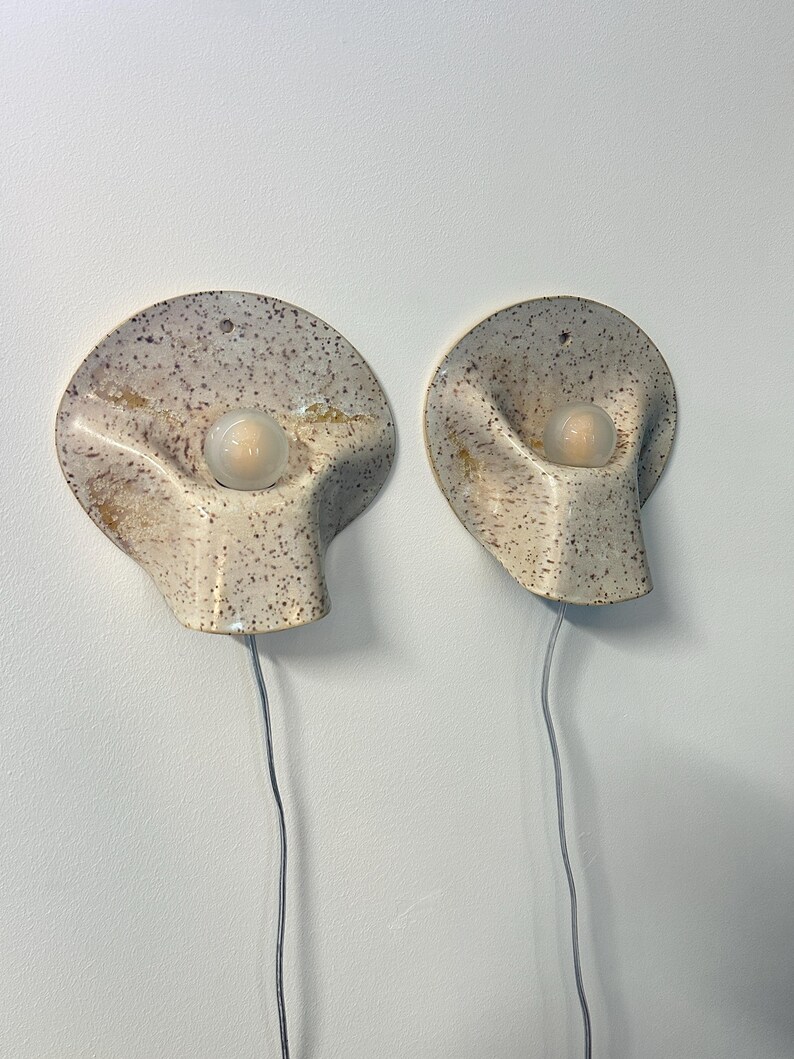 May include: A pair of wall-mounted sconces featuring a speckled cream ceramic shell-like design with a central frosted glass bulb. Each fixture has a curved, organic shape with a small hole at the top for mounting and a thin grey electrical cord.
