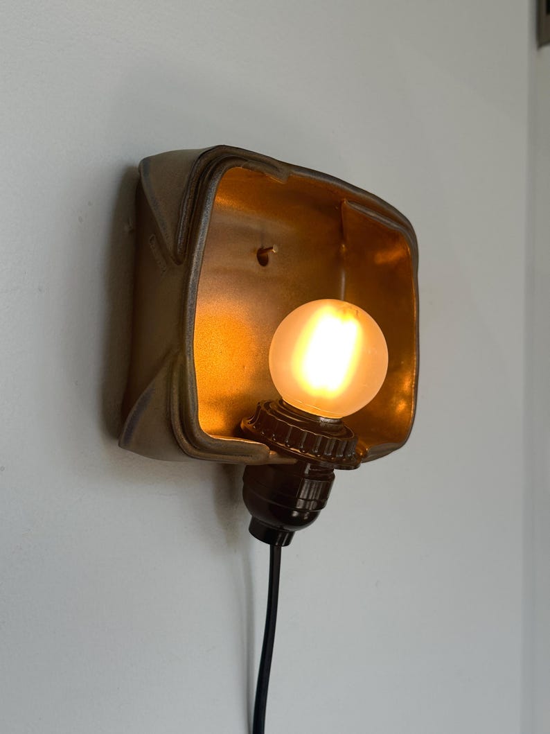 May include: A vintage-style wall sconce with a warm, glowing light bulb. The sconce has a square, metallic frame with a gold interior and a dark brown base. The light bulb is round and emits a soft, yellow light. The sconce is mounted on a white wall.