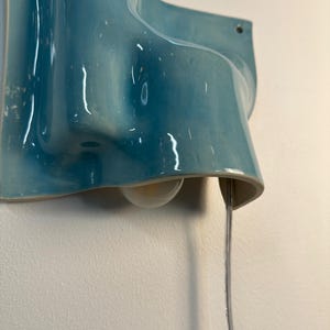 May include: A wall-mounted light fitting featuring a glossy, wavy ceramic shade in a vibrant turquoise blue glaze. The fitting includes a single exposed light bulb tucked beneath the curved ceramic form and a clear electrical cord hanging downwards.
