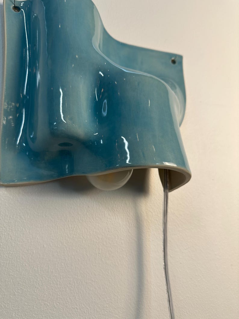 May include: A wall-mounted light fitting featuring a glossy, wavy ceramic shade in a vibrant turquoise blue glaze. The fitting includes a single exposed light bulb tucked beneath the curved ceramic form and a clear electrical cord hanging downwards.