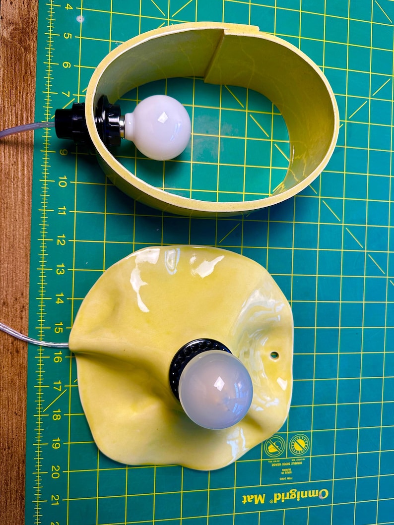 May include: Two yellow light fixtures with white bulbs and black sockets. The top fixture is an oval band, while the bottom fixture has a wavy, organic shape with a small circular hole near the edge. Both include clear electrical cords.
