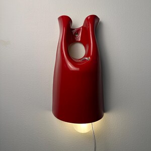May include: A glossy red wall-mounted lamp with a smooth, curved, bell-like silhouette. The fixture features a circular cutout near the top and a single light bulb emitting a warm glow from the bottom, connected by a thin, clear electrical cord.