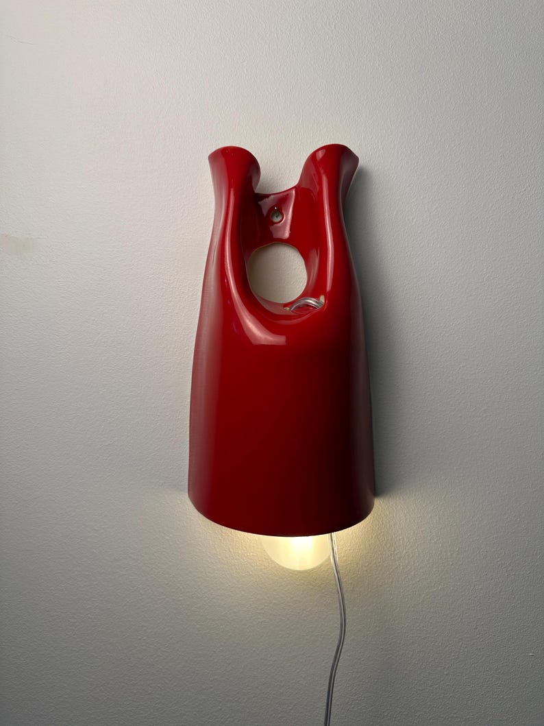 May include: A glossy red wall-mounted lamp with a smooth, curved, bell-like silhouette. The fixture features a circular cutout near the top and a single light bulb emitting a warm glow from the bottom, connected by a thin, clear electrical cord.