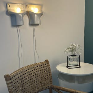 May include: Two light gray ceramic wall sconces with glowing bulbs, each featuring a white power cord. A woven rattan chair sits in the foreground, while a small square glass vase with white flowers rests on a round white side table nearby.