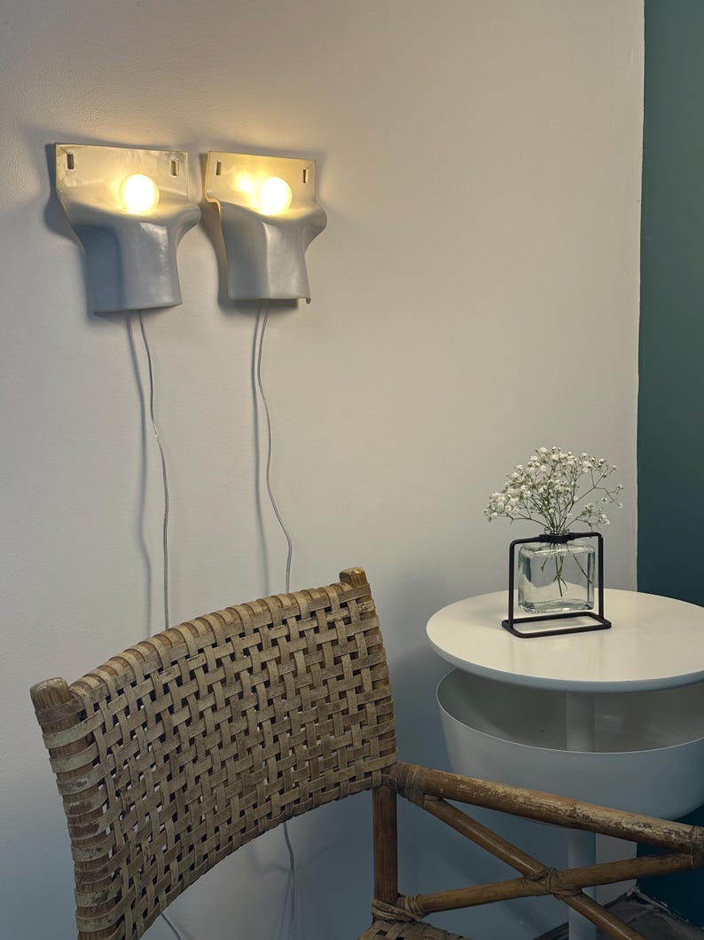 May include: Two light gray ceramic wall sconces with glowing bulbs, each featuring a white power cord. A woven rattan chair sits in the foreground, while a small square glass vase with white flowers rests on a round white side table nearby.