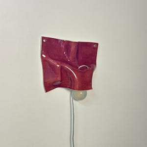 May include: A wall-mounted ceramic light fixture with a glossy, deep red, undulating surface. The piece features two circular mounting holes at the top corners and a central opening holding a round, frosted glass light bulb with a white power cord.
