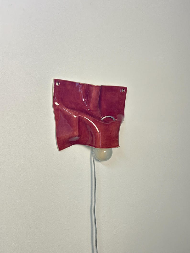 May include: A wall-mounted ceramic light fixture with a glossy, deep red, undulating surface. The piece features two circular mounting holes at the top corners and a central opening holding a round, frosted glass light bulb with a white power cord.