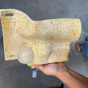 May include: A wall-mounted ceramic light fixture with a yellow glaze and dark brown speckles. The piece features an irregular, organic shape with a smooth surface and a single round, frosted glass light bulb protruding from the front face.