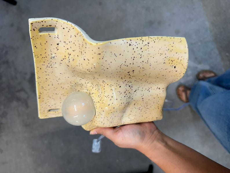 May include: A wall-mounted ceramic light fixture with a yellow glaze and dark brown speckles. The piece features an irregular, organic shape with a smooth surface and a single round, frosted glass light bulb protruding from the front face.