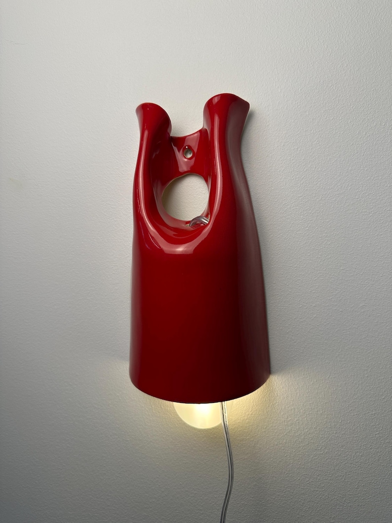 May include: A glossy red wall-mounted lamp with a smooth, curved, elongated form featuring a central circular cutout near the top. The fixture has a wide, open base that emits a warm, soft light from a single bulb, with a clear power cord extending downward.