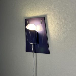 Pode incluir: Uma moderna lumin&aacute;ria de parede em azul escuro. Possui uma sombra curva e uma l&acirc;mpada redonda e brilhante. Um cabo de alimenta&ccedil;&atilde;o prateado pende. A lumin&aacute;ria &eacute; fixada a uma placa retangular azul escuro.
