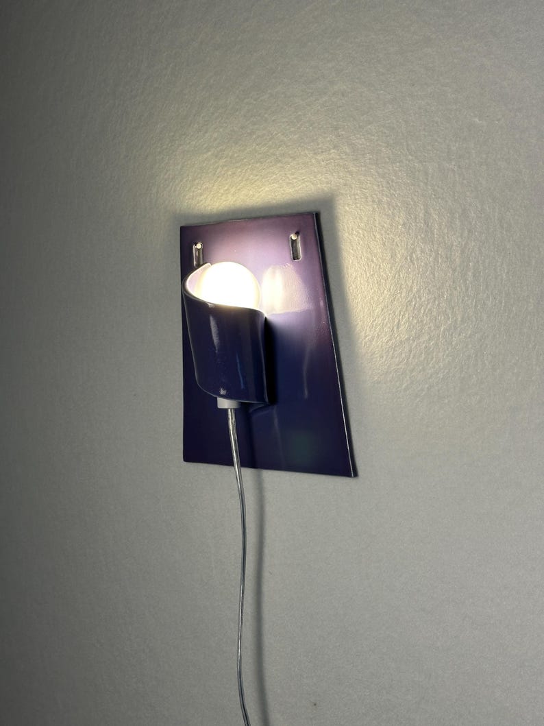 Pode incluir: Uma moderna lumin&aacute;ria de parede em azul escuro. Possui uma sombra curva e uma l&acirc;mpada redonda e brilhante. Um cabo de alimenta&ccedil;&atilde;o prateado pende. A lumin&aacute;ria &eacute; fixada a uma placa retangular azul escuro.