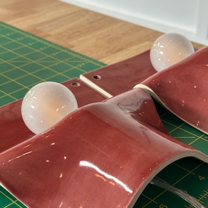 May include: Two wall sconces featuring curved, glossy, deep red ceramic shades and round, white glass light bulbs. Each fixture includes a small circular mounting hole at the top edge. The sconces are positioned on a green cutting mat with visible inch markings.
