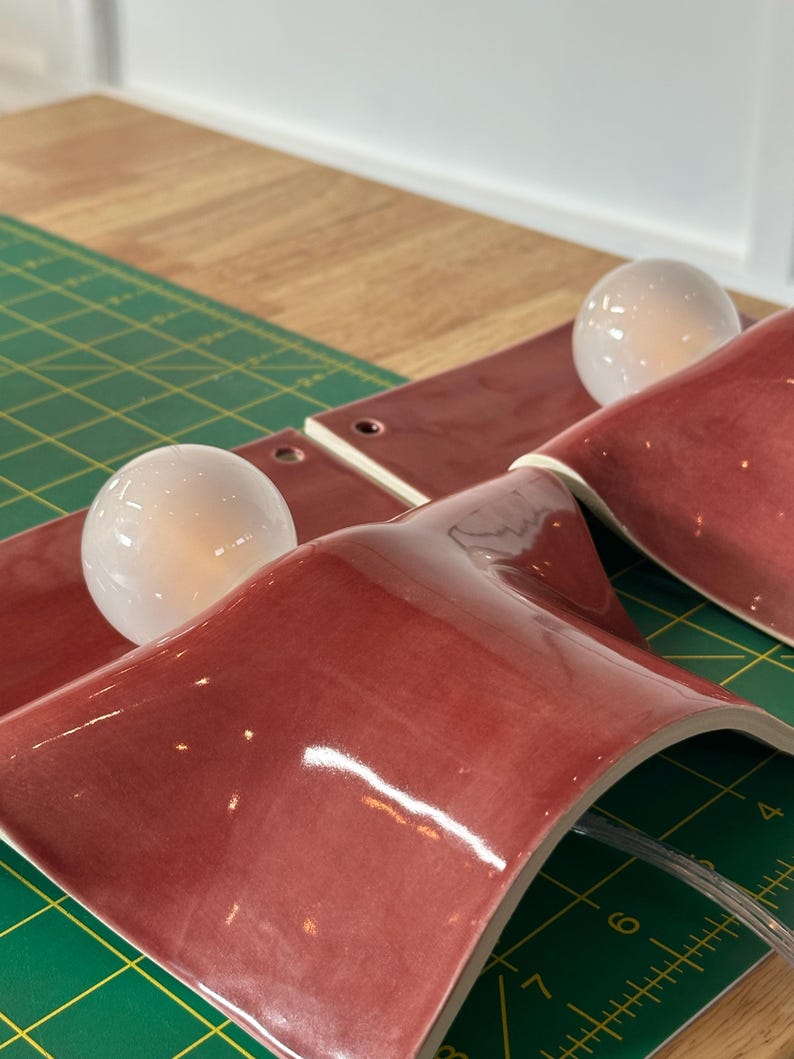 May include: Two wall sconces featuring curved, glossy, deep red ceramic shades and round, white glass light bulbs. Each fixture includes a small circular mounting hole at the top edge. The sconces are positioned on a green cutting mat with visible inch markings.