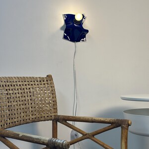 May include: A wall-mounted lamp featuring a glossy, deep blue, irregularly shaped ceramic or metal shade with a single glowing light bulb. A thin, white electrical cord hangs vertically from the fixture down the wall toward the floor.