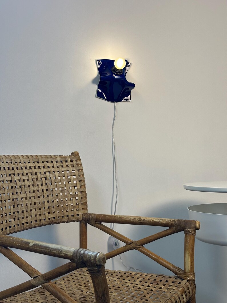 May include: A wall-mounted lamp featuring a glossy, deep blue, irregularly shaped ceramic or metal shade with a single glowing light bulb. A thin, white electrical cord hangs vertically from the fixture down the wall toward the floor.