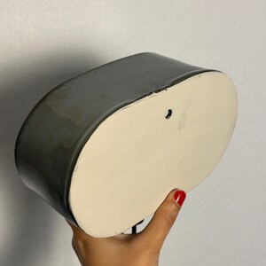 K&ouml;nnte beinhalten: Ein grauer Wandtopf aus Keramik mit glatter, gl&auml;nzender Oberfl&auml;che. Der Pflanztopf hat eine ovale Form mit einer flachen, cremefarbenen Basis und einem Loch zum Aufh&auml;ngen. Der Pflanztopf wird von einer Person gehalten.
