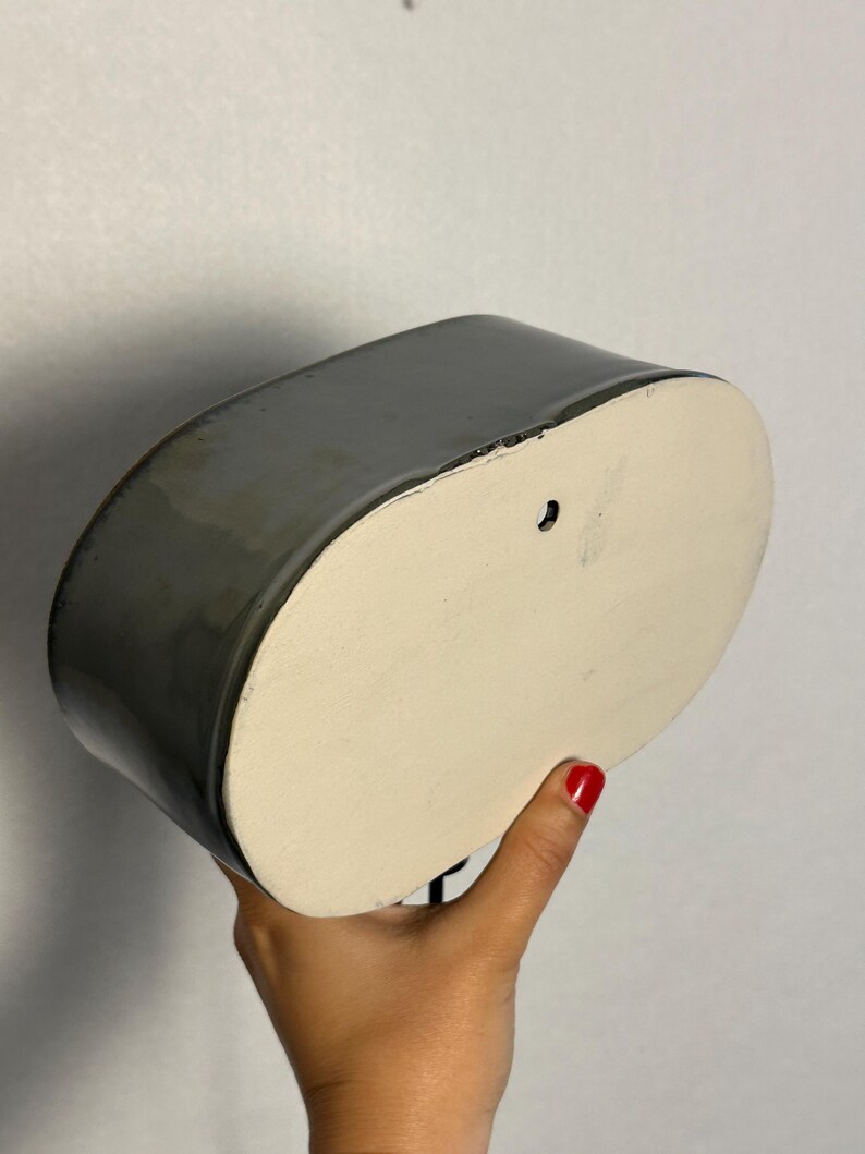 K&ouml;nnte beinhalten: Ein grauer Wandtopf aus Keramik mit glatter, gl&auml;nzender Oberfl&auml;che. Der Pflanztopf hat eine ovale Form mit einer flachen, cremefarbenen Basis und einem Loch zum Aufh&auml;ngen. Der Pflanztopf wird von einer Person gehalten.