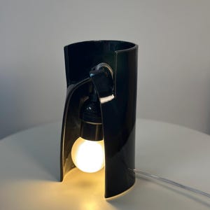 May include: A glossy black ceramic table lamp with a curved, cylindrical structure that is open at the front. A single round, glowing white light bulb is suspended inside the dark frame, connected by a black socket and a thin, clear power cord.
