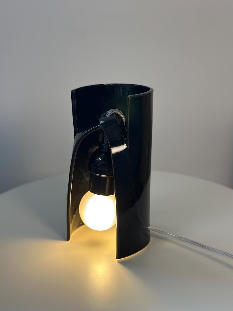 May include: A glossy black ceramic table lamp with a curved, cylindrical structure that is open at the front. A single round, glowing white light bulb is suspended inside the dark frame, connected by a black socket and a thin, clear power cord.
