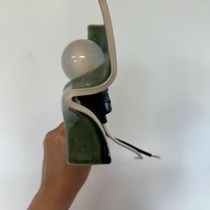 May include: A wall sconce featuring a curved, glossy olive green ceramic shade with a smooth finish. The fixture includes a round, frosted glass light bulb seated in a black socket, with exposed white and black electrical wires extending outward.
