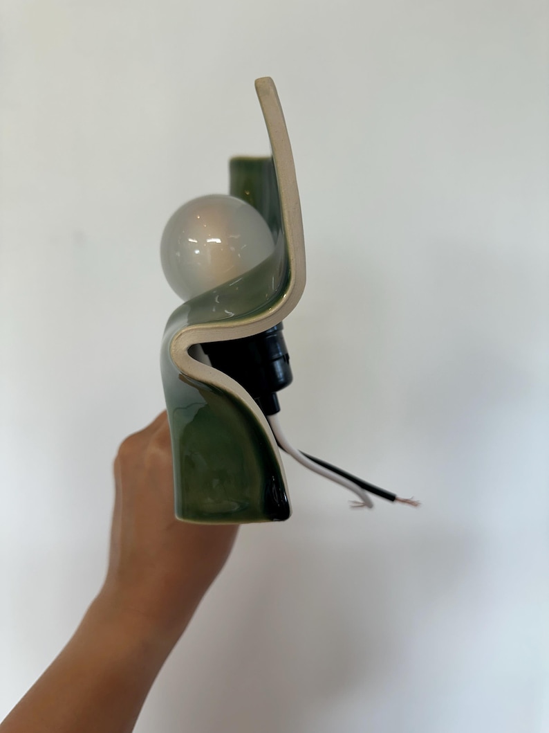 May include: A wall sconce featuring a curved, glossy olive green ceramic shade with a smooth finish. The fixture includes a round, frosted glass light bulb seated in a black socket, with exposed white and black electrical wires extending outward.