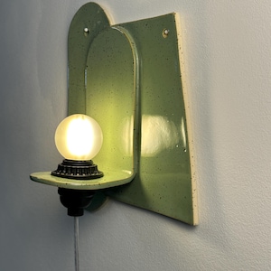 May include: A wall-mounted light fixture featuring a glossy, light green ceramic backplate with dark speckles. The fixture includes a small, horizontal shelf holding a black socket and a glowing, frosted spherical light bulb with a clear power cord.