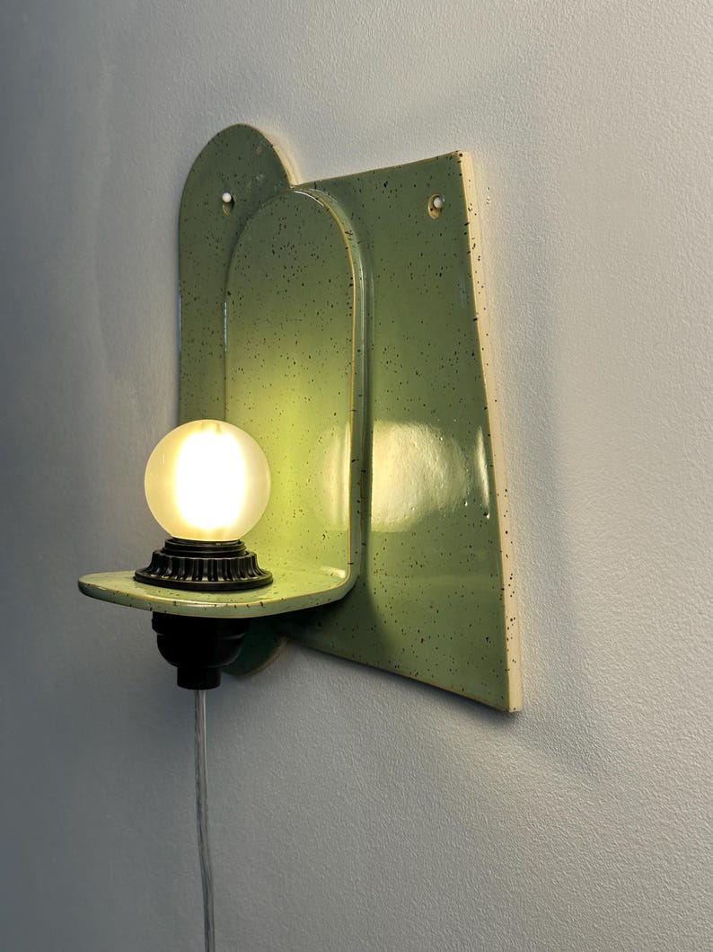 May include: A wall-mounted light fixture featuring a glossy, light green ceramic backplate with dark speckles. The fixture includes a small, horizontal shelf holding a black socket and a glowing, frosted spherical light bulb with a clear power cord.