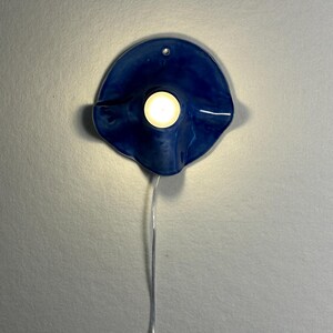 May include: A blue ceramic wall sconce with a circular design. The sconce has a small hole at the top for hanging and a light in the center. A thin, silver wire extends from the bottom of the sconce.
