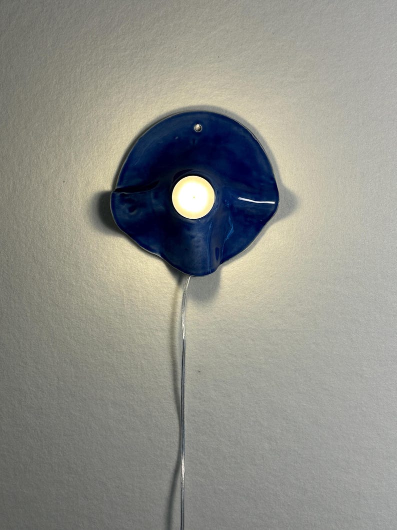 May include: A blue ceramic wall sconce with a circular design. The sconce has a small hole at the top for hanging and a light in the center. A thin, silver wire extends from the bottom of the sconce.