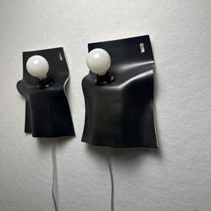 May include: Two wall-mounted lamps featuring a matte black, curved rectangular shade that drapes over a central light socket. Each lamp includes a round, white frosted light bulb and a thin grey power cord extending from the bottom of the fixture.