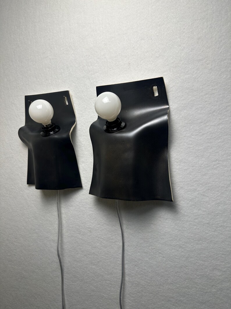 May include: Two wall-mounted lamps featuring a matte black, curved rectangular shade that drapes over a central light socket. Each lamp includes a round, white frosted light bulb and a thin grey power cord extending from the bottom of the fixture.