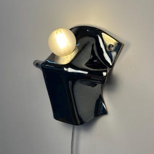 May include: A wall-mounted lamp featuring a glossy black ceramic base with an irregular, fluid shape. A single round, frosted glass light bulb is set into the top of the fixture, emitting a warm glow. A thin power cord hangs from the bottom.