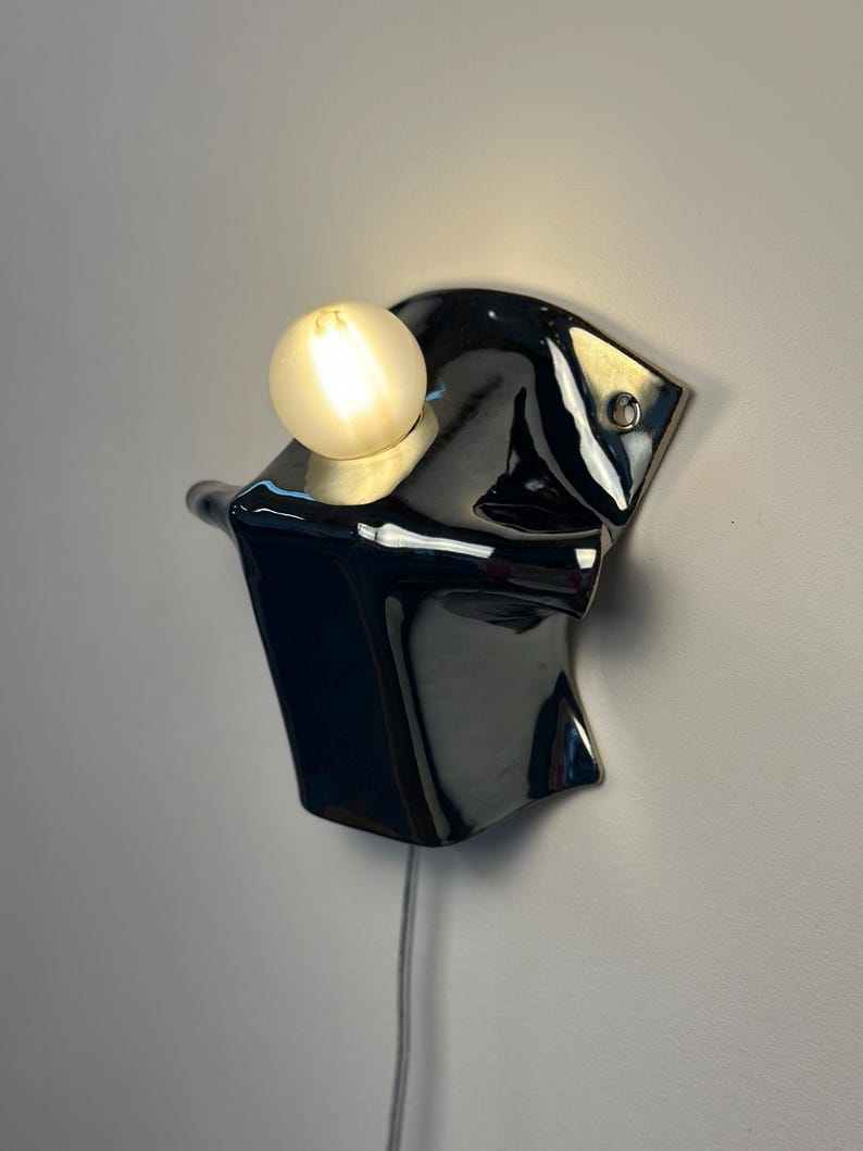 May include: A wall-mounted lamp featuring a glossy black ceramic base with an irregular, fluid shape. A single round, frosted glass light bulb is set into the top of the fixture, emitting a warm glow. A thin power cord hangs from the bottom.