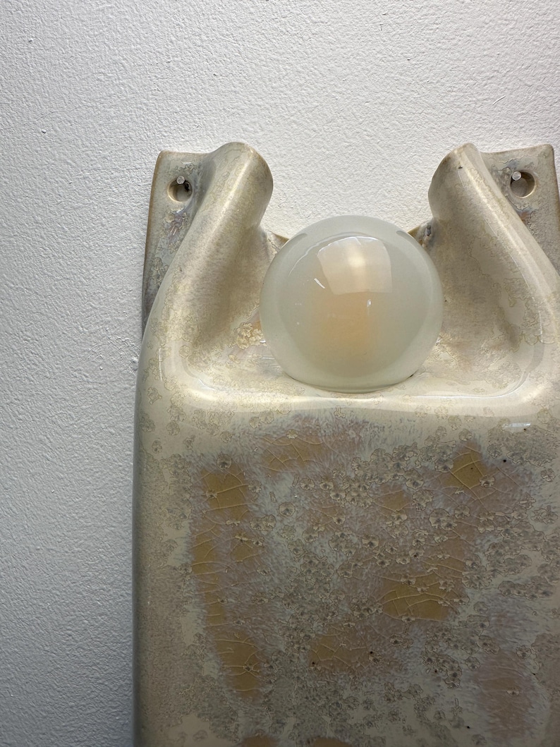 May include: A wall-mounted ceramic light fitting featuring a glossy, cream-coloured glaze with a crackled texture and golden-brown crystalline patterns. The fitting has a sculptural, organic shape with two mounting holes and a central frosted glass globe.