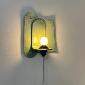 May include: A wall-mounted light fixture featuring a pale green, speckled ceramic backplate with an arched design. A small, round, glowing light bulb sits on a horizontal shelf extending from the center, connected to a black base and a white cord.